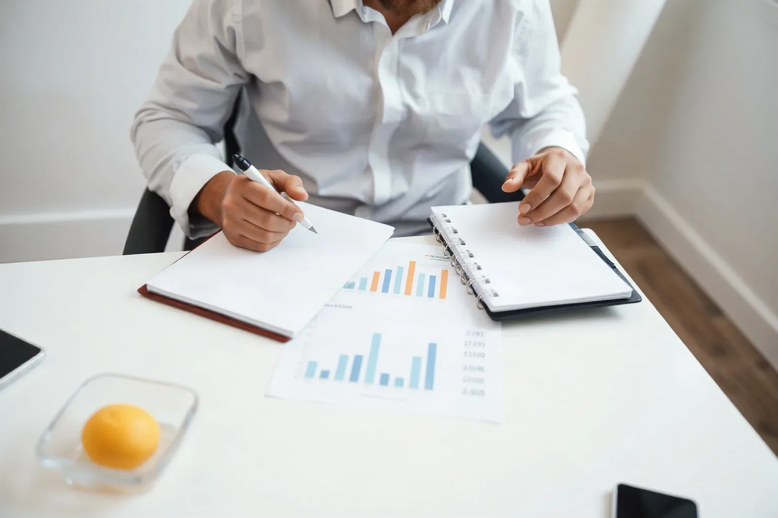 A marketing professional seated at a focused desk workspace laying two ring-bound notebooks open side by side and comparing hand-annotated pages while a printed bar-chart report is spread at the centre of the desk