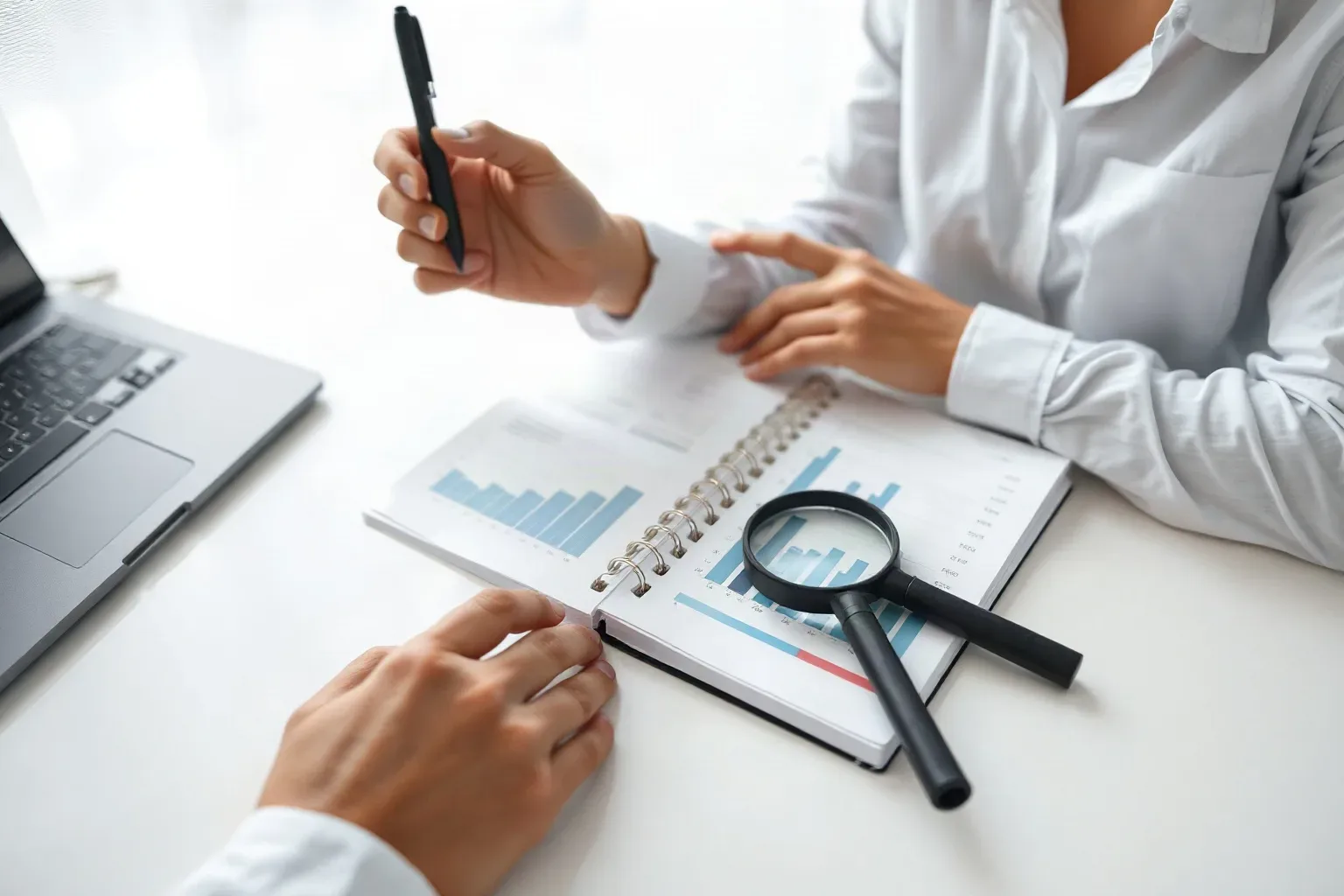 A marketing professional at a focused single-desk workspace flipping to a tabbed section of a ring-bound notebook and ticking off on-page elements while a printed analytics report with bar charts is anchored open beside it with the magnifying glass